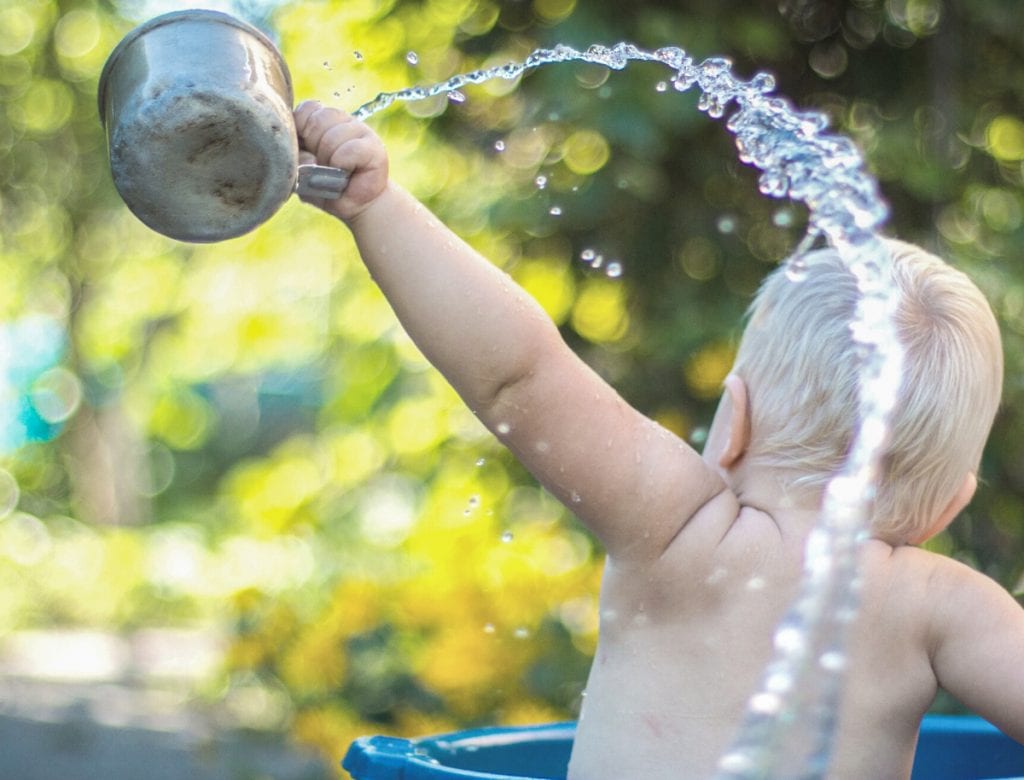 Baby spritzt Wasser, Aufnahme von hinten in Badwanne mit Becher in der Hand