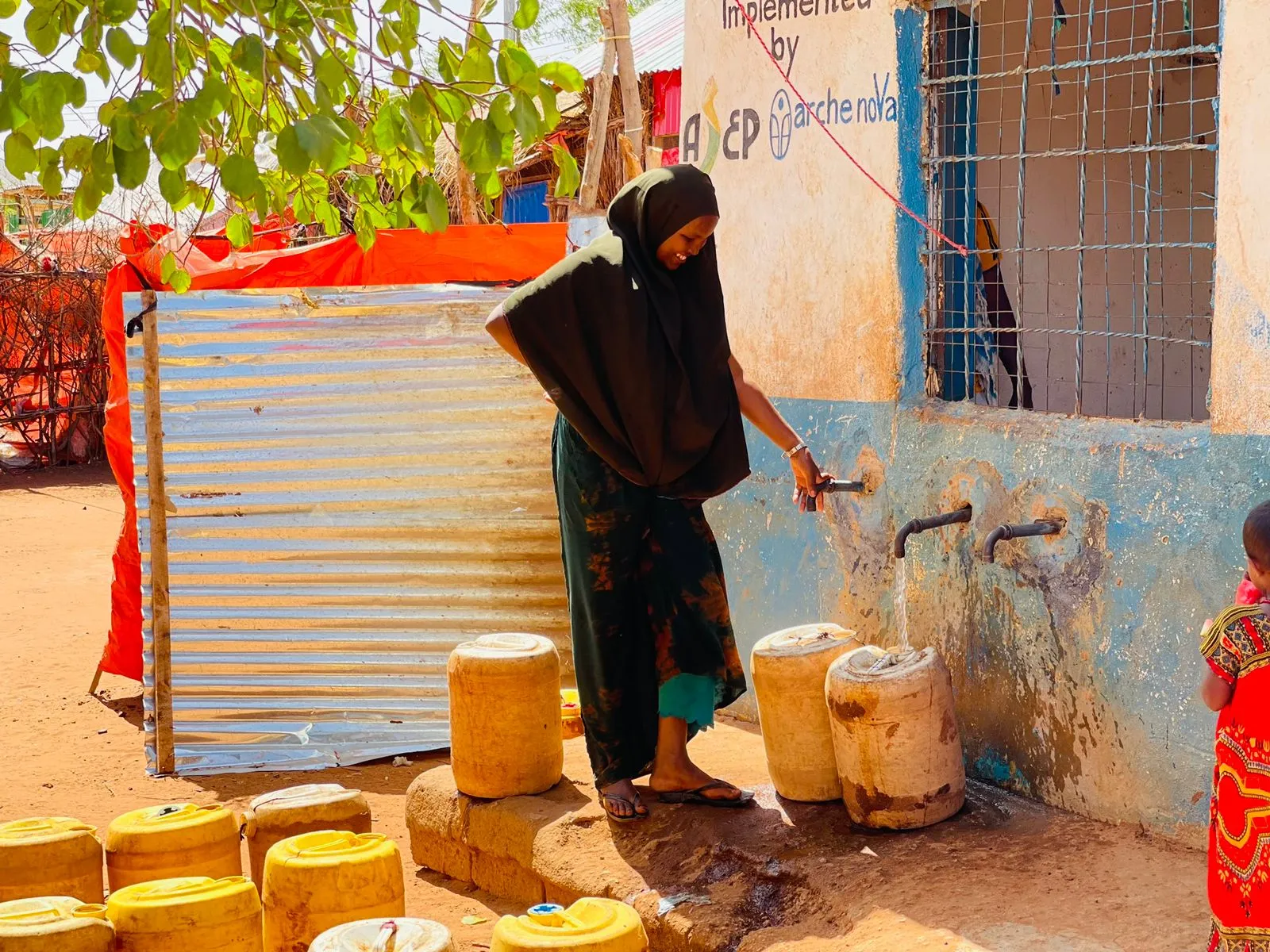 Eine Frau füllt gelbe Wasserkanister an einem Wasserhahn in einem ländlichen Gebiet ab.