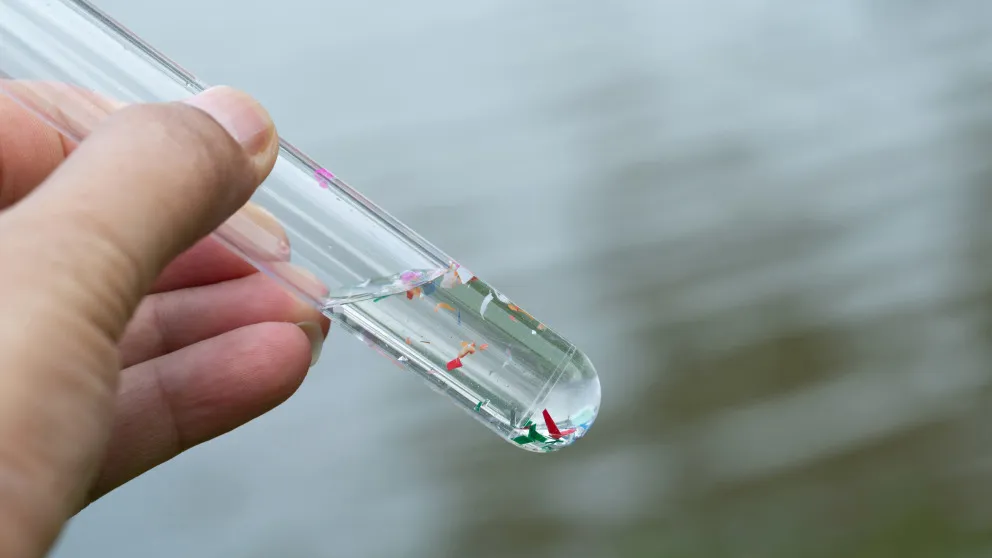 Kunststoffpartikel im Trinkwasser sind längst Realität Hand hält ein Reagenzglas mit Wasserprobe, darin schwimmen bunte Mikroplastikpartikel.
