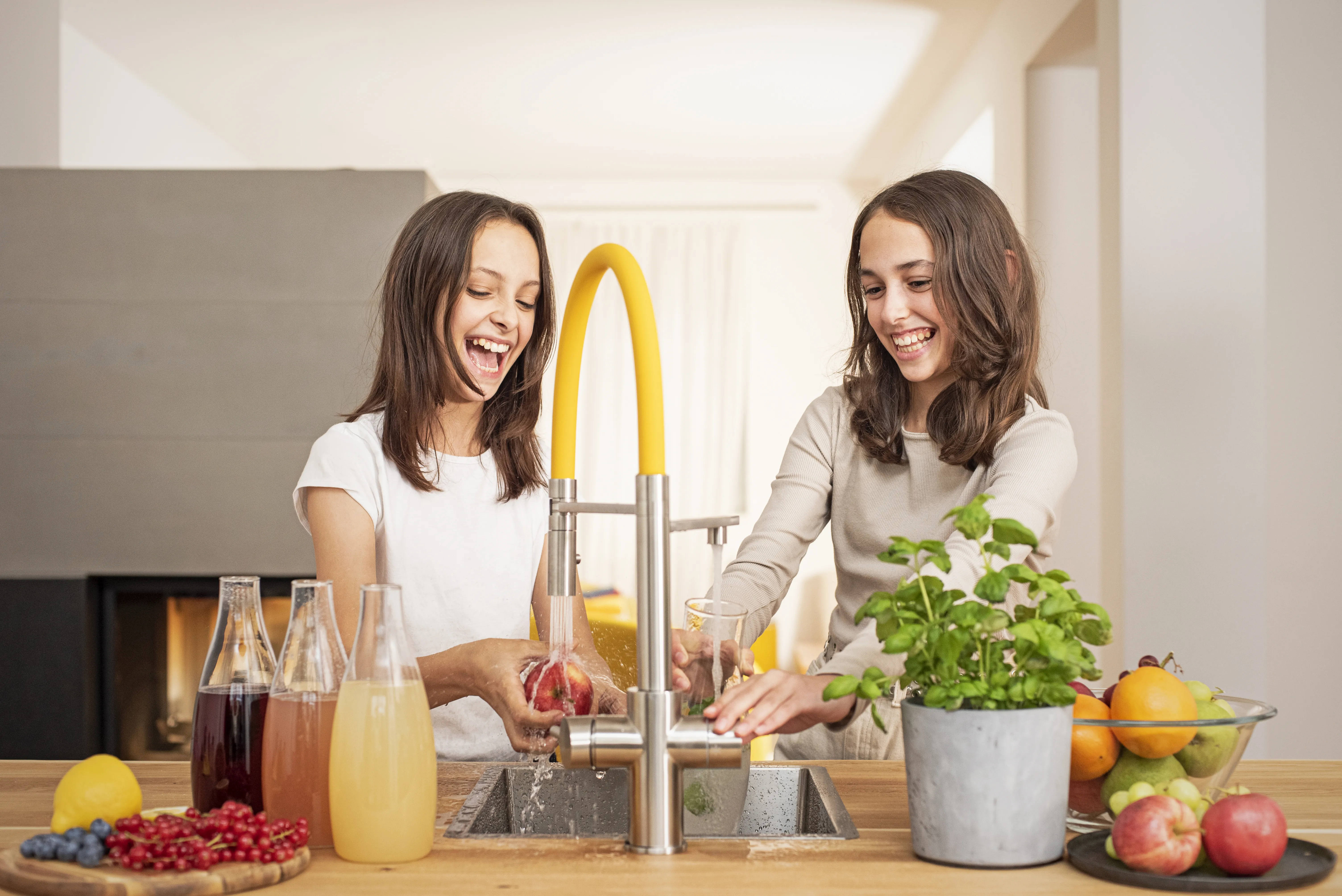Wasserfilterung mit dem Living Water Wasserhahn schützt vor Nanoplastik im Alltag Zwei Mädchen lachen in der Küche und nutzen gemeinsam den Living Water Wasserhahn, daneben stehen Früchte und Karaffen.