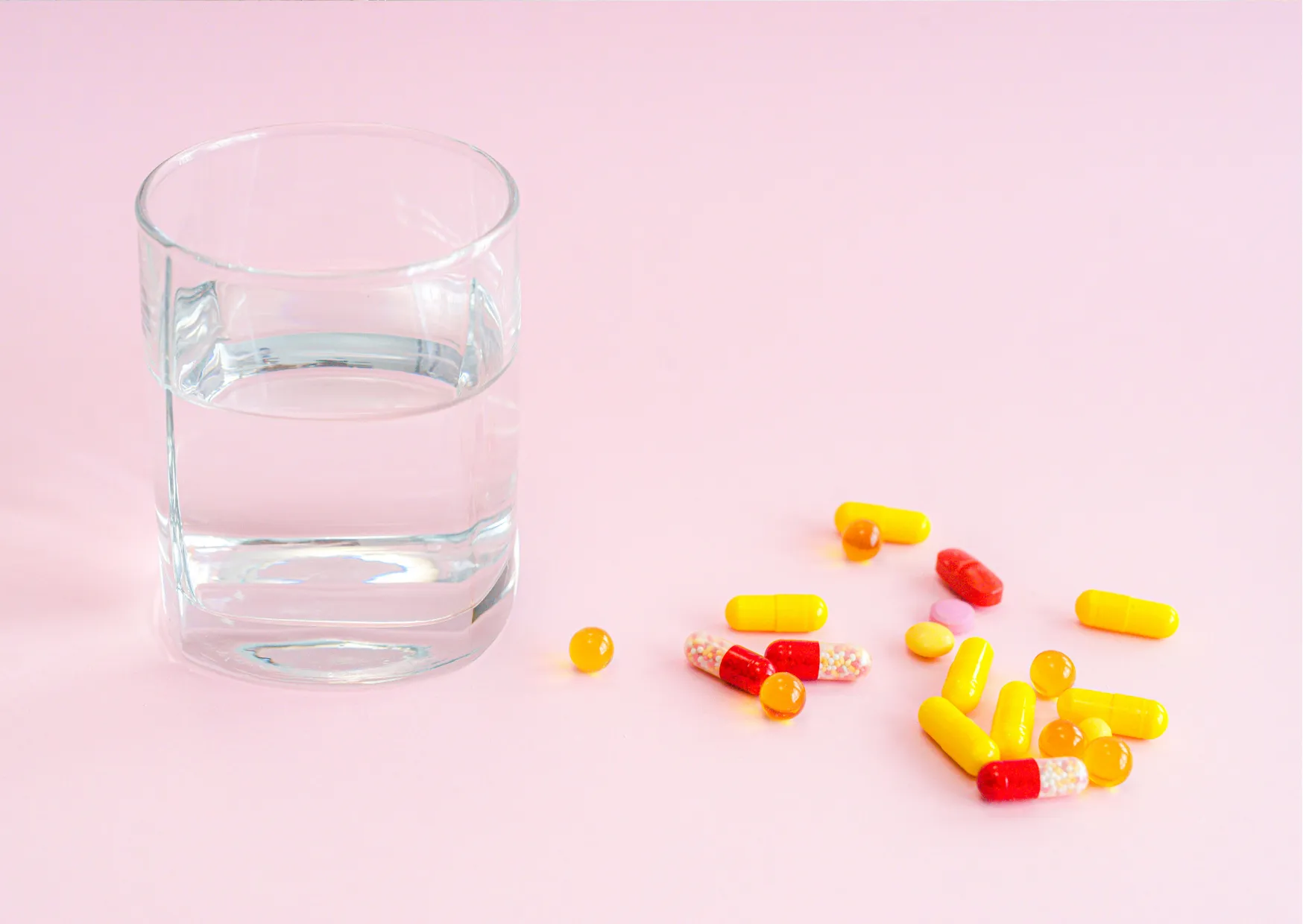 Glas mit Wasser neben bunten Medikamentenkapseln auf rosa Hintergrund