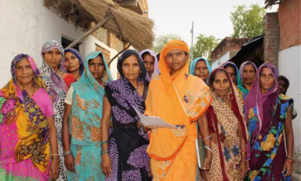 Gruppierung von Frauen in bunten traditionellen Kleidern, die zusammen stehen und in die Kamera schauen. Eine Frau in orange hebt ein Dokument in der Hand.