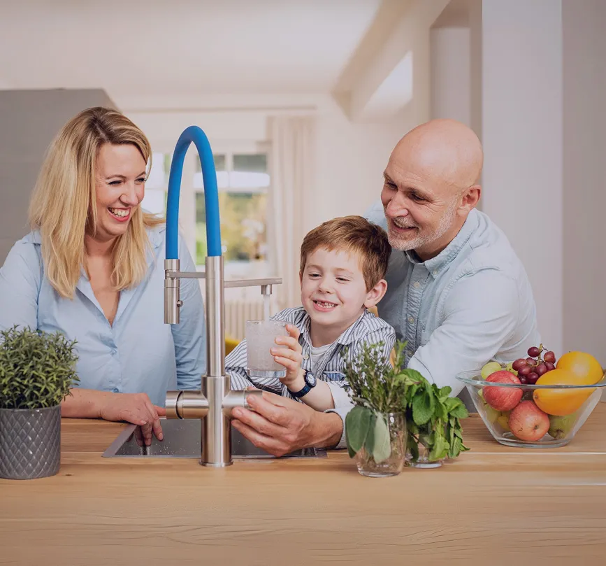 Eine Familie steht an der Küchentheke, lächelt und hat Spaß. Ein Junge hält ein Glas Wasser, während die Frau und der Mann ihm helfen. Frische Kräuter und Obst sind sichtbar.