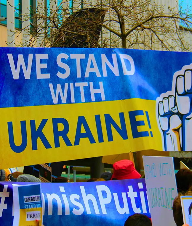 Demonstration für die Ukraine mit einem großen Banner, auf dem "WE STAND WITH UKRAINE!" geschrieben steht, und weiteren Schildern im Hintergrund.