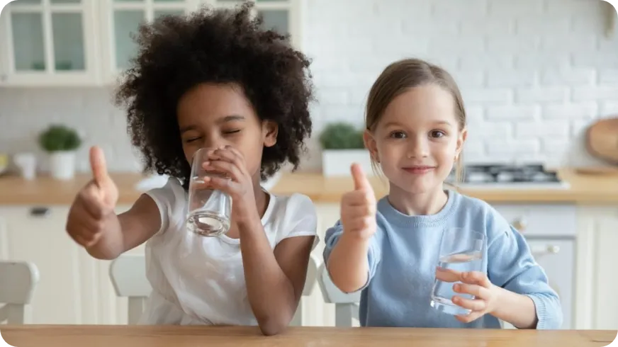 Kinder trinken gerne Wasser und haben Spaß dabei Zwei Kinder sitzen in der Küche und trinken Wasser, beide zeigen Daumen hoch.