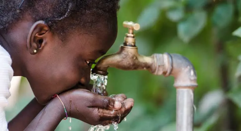 Ein Mädchen beugt sich über einen Wasserhahn und trinkt mit auf Händen gefasstem Wasser. Grüne Pflanzen im Hintergrund.